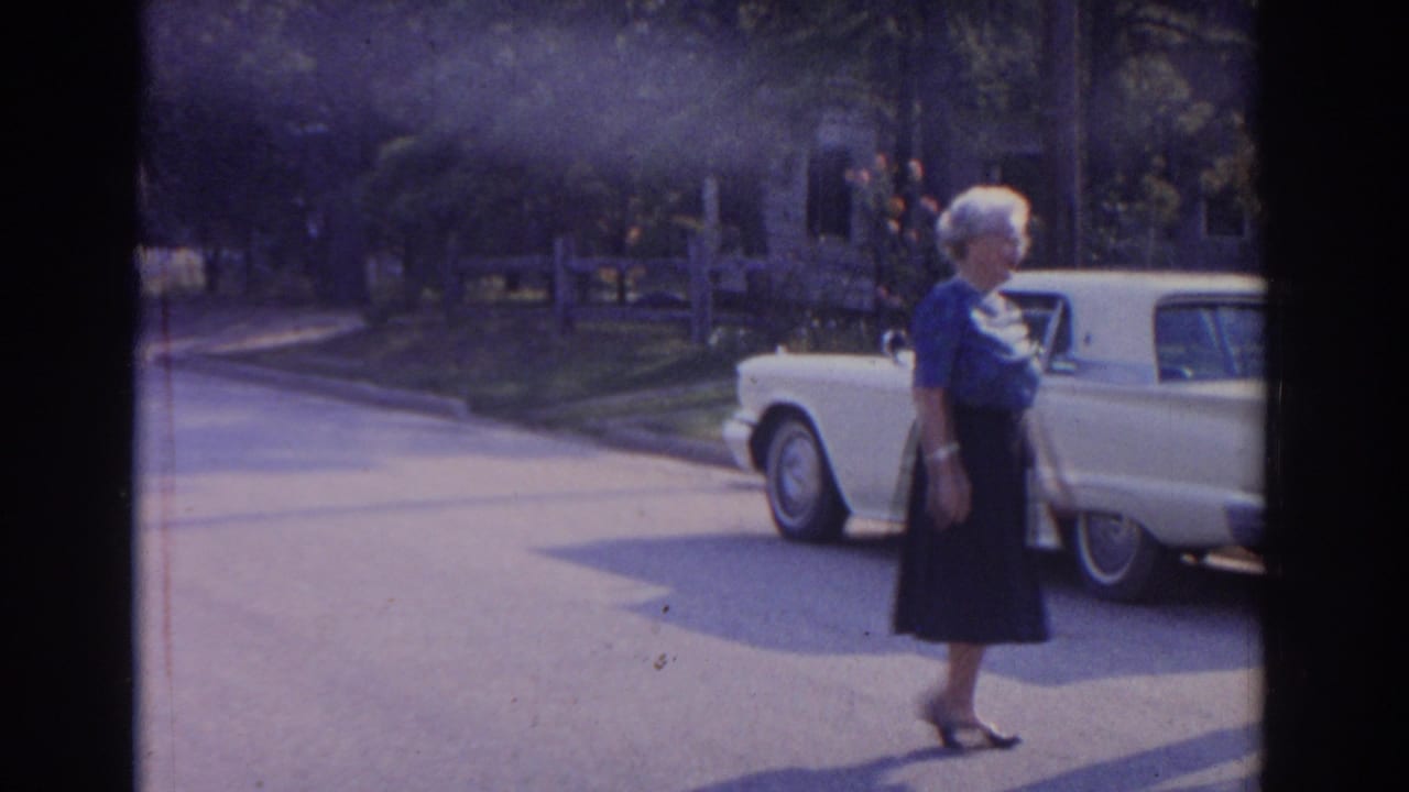 Older woman in blue top walking past a white classic car on the street.