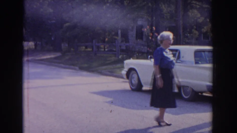 1963 New Jersey home-movie frame with an older woman walking in front of a parked white classic car in a residential neighborhood.