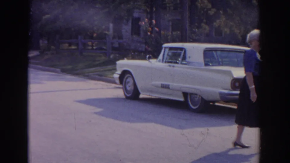 1963 New Jersey home-movie frame showing a parked white classic car beside the curb with an older woman partly visible at right.