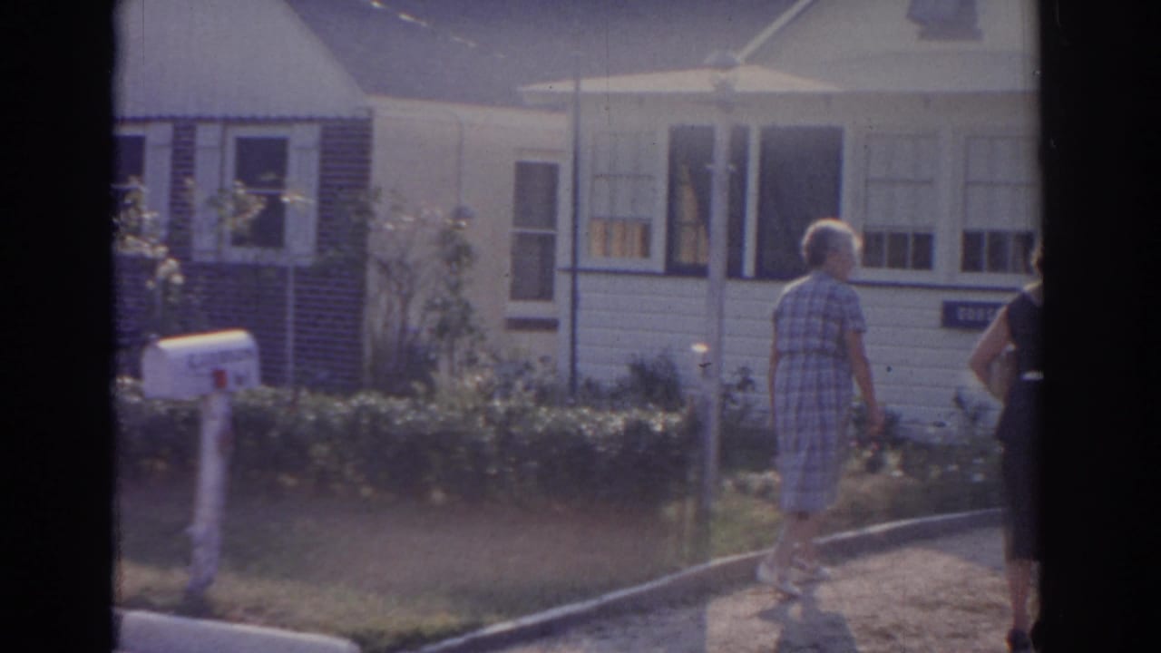 Woman in plaid dress walks up a driveway toward a white-sided suburban house.