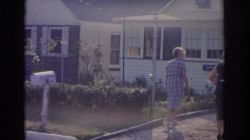 In 1963 New Jersey home-movie footage, a woman in a plaid dress walks along a driveway toward a white-sided house with a mailbox nearby.