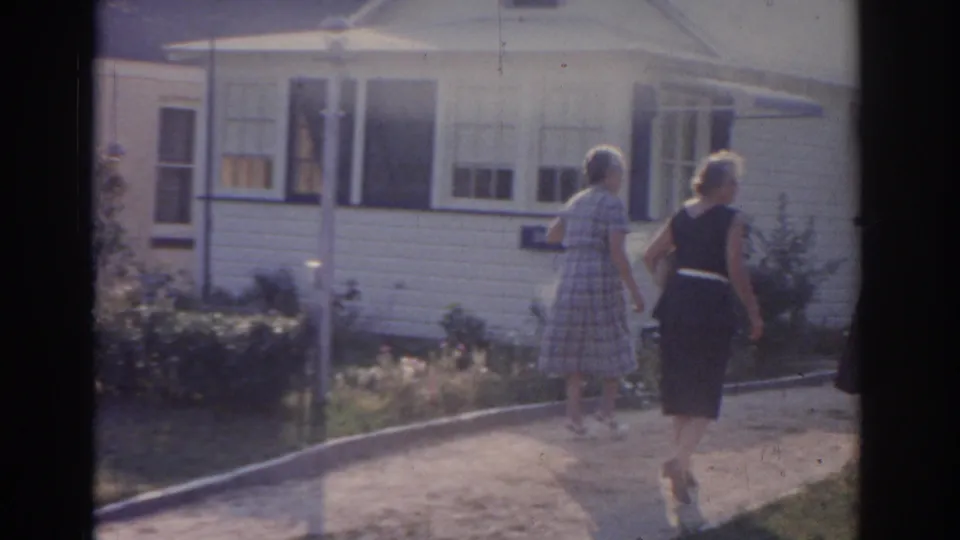 Two women walk along a suburban driveway in New Jersey, 1963, with a white-sided house, front windows, shrubs, and mailbox in view.