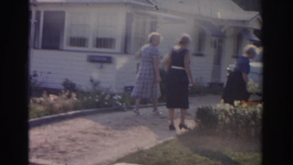 Three women appear near the driveway and garden of a white-sided New Jersey suburban house, with 1960s home-movie softness and grain.