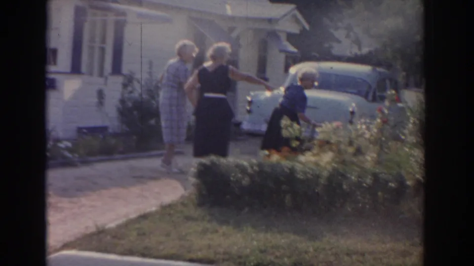 Three women gather near a driveway and white period car outside a New Jersey suburban home in 1963 home-movie footage.