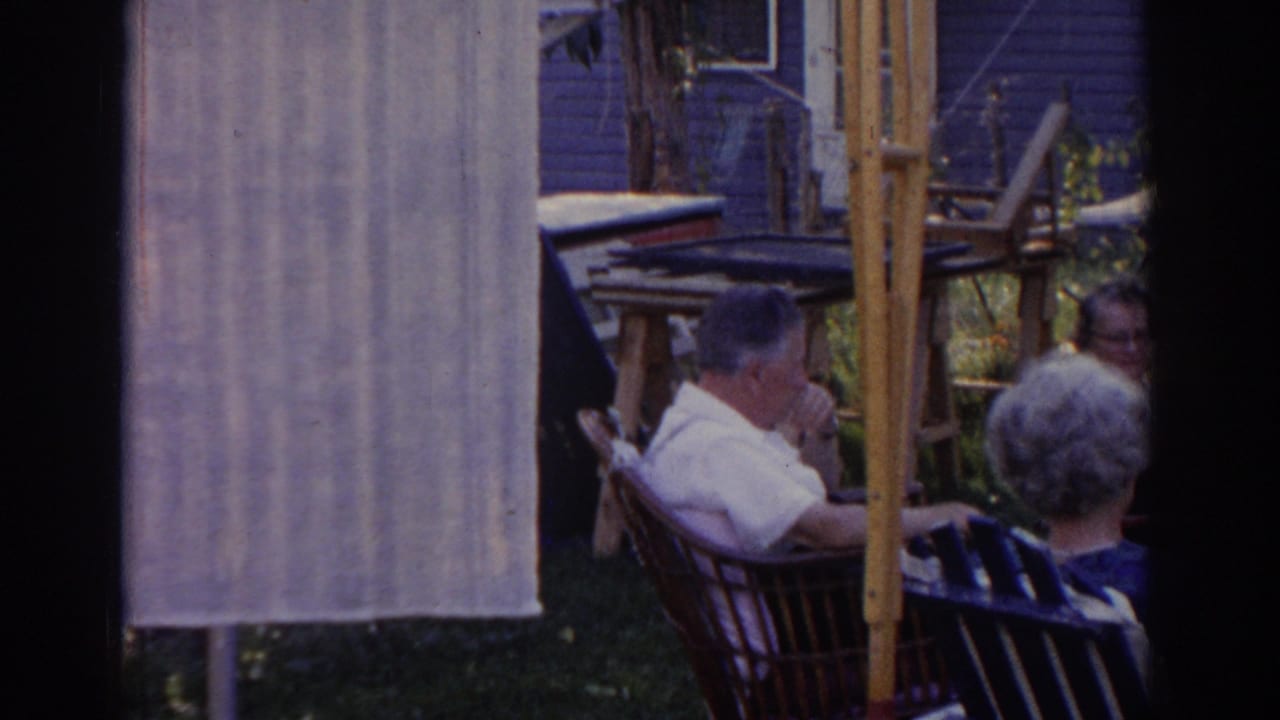 1963 New Jersey home-movie frame of older adults seated in a backyard, partly framed by film gate shadow and a pale hanging cloth.