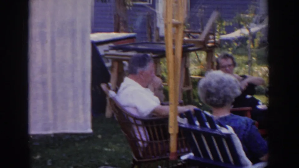 1963 New Jersey backyard scene with older adults in wicker and folding chairs, a purple-sided house, and sunlit plants in the background.