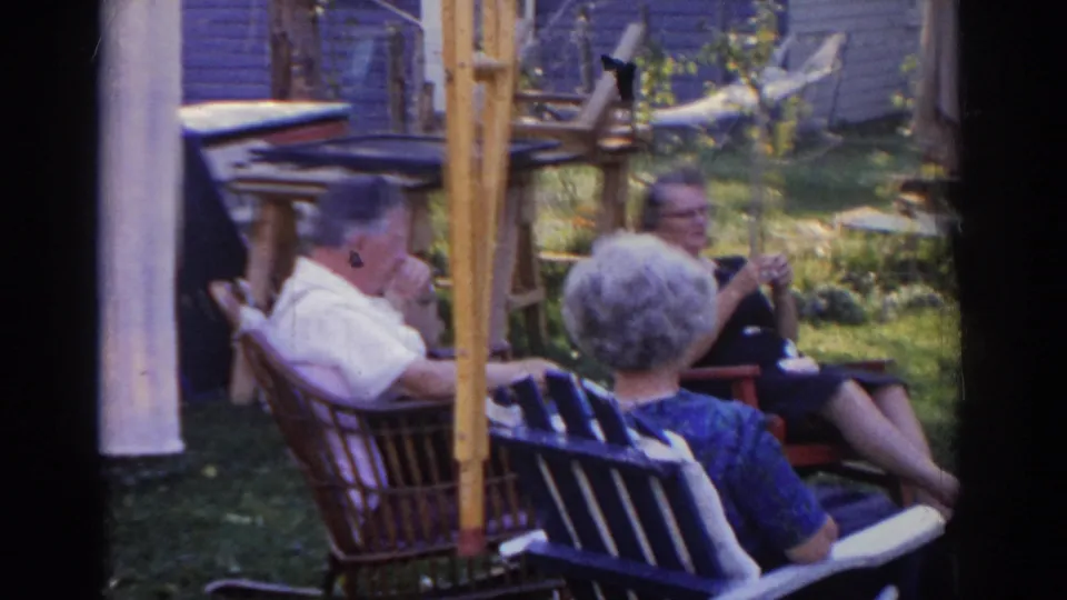 Home-movie frame from 1963 New Jersey showing older adults seated in a grassy backyard, with folding chairs, a house wall, and yard structures behind them.