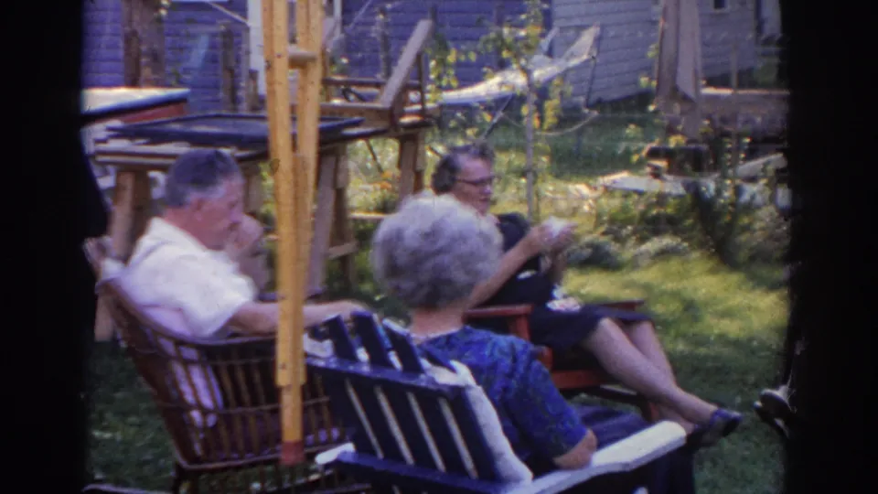 1963 New Jersey 8mm frame of older adults relaxing in a backyard conversation, with a folding chair, wicker chair, purple house siding, and sunlit garden details.