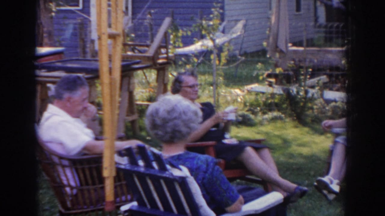 In 1963 New Jersey home-movie footage, three older adults sit in lawn chairs in a backyard near a house and garden.