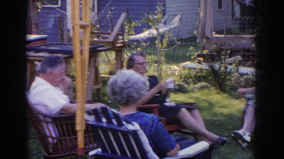 In 1963 New Jersey home-movie footage, three older adults sit in lawn chairs in a backyard near a house and garden.