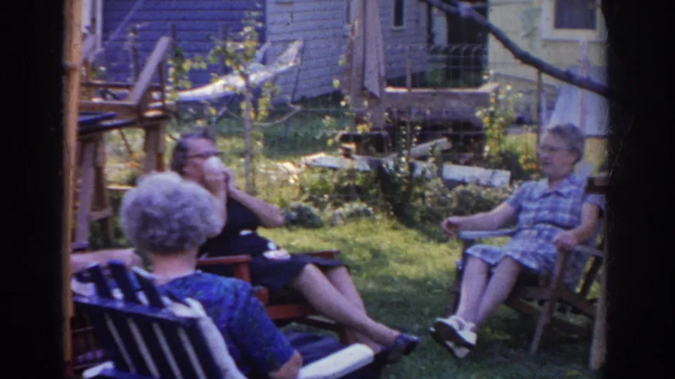 A woman in a plaid dress is more visible as three older adults sit facing one another in the 1963 backyard scene.
