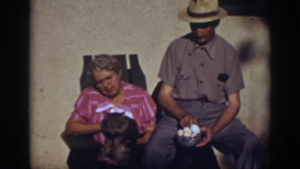 Auburn home-movie frame showing the seated woman cradling a dark cat as the man in a brimmed hat holds a bowl containing white eggs.