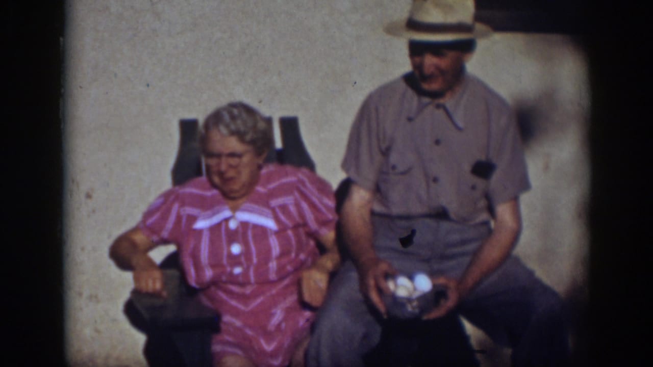 Older couple seated outdoors, with the woman facing camera and the man holding eggs beside her.