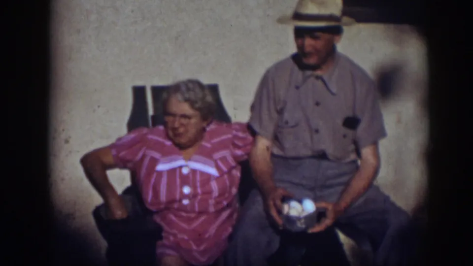 A 1939 Auburn home-movie frame shows the woman leaning in her chair while the man remains beside her with eggs in his lap.