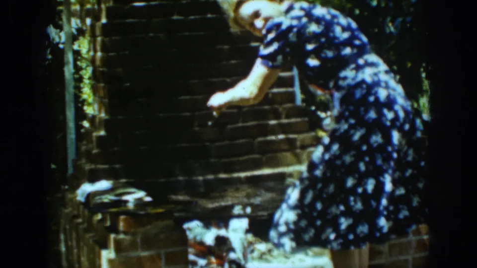 A woman in a blue patterned dress bends near a brick hearth in the 1939 Auburn, California home-movie scene.