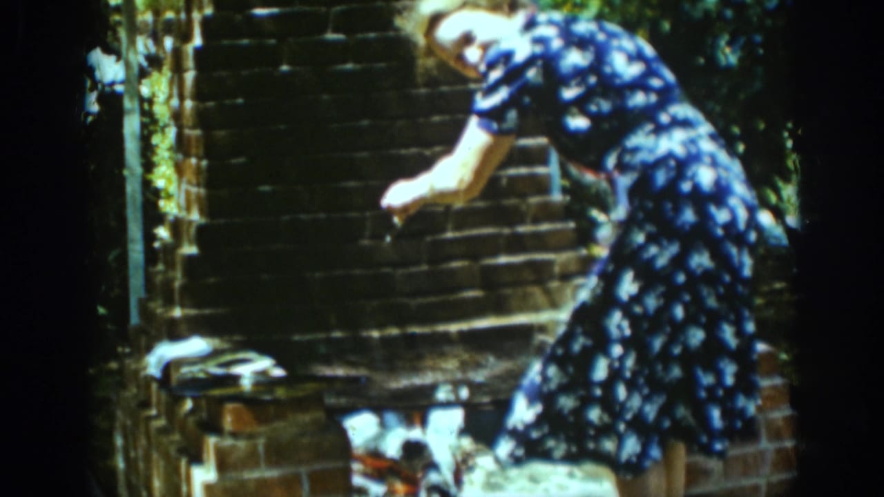 Woman in a blue floral dress tends food at a brick outdoor fireplace.