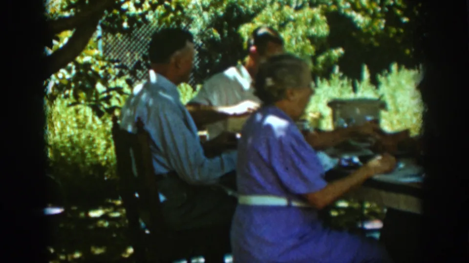 A 1939 Auburn backyard meal scene shows adults seated close together at a table, partly shaded by trees and surrounded by garden foliage.