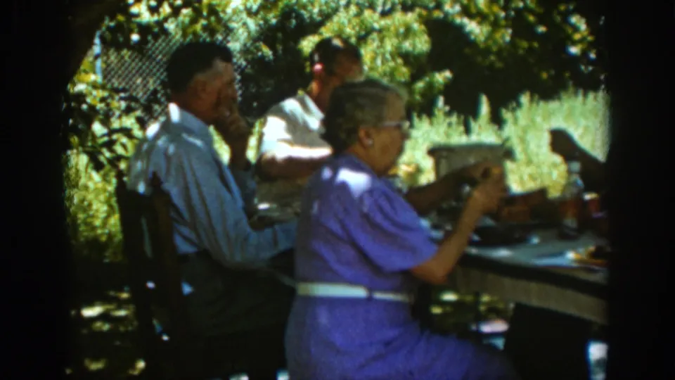 Adults at a shaded backyard table in Auburn, California, 1939, with dishes and bottles on the table and foliage behind them.