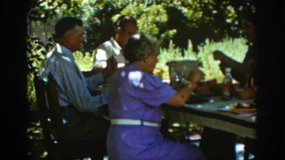 In Auburn, California in 1939, adults sit at a shaded outdoor table with plates, food, and bottles during a family meal.