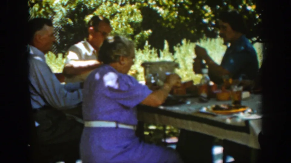 A 1939 Auburn home-movie frame shows adults seated around a table under trees, with food, bottles, and tableware visible.