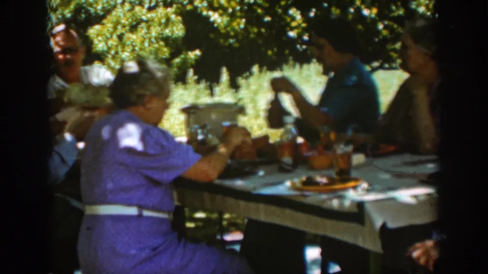 The Auburn, California home movie shows a woman in purple seated at a shaded meal table with adults, plates, bottles, and food nearby.