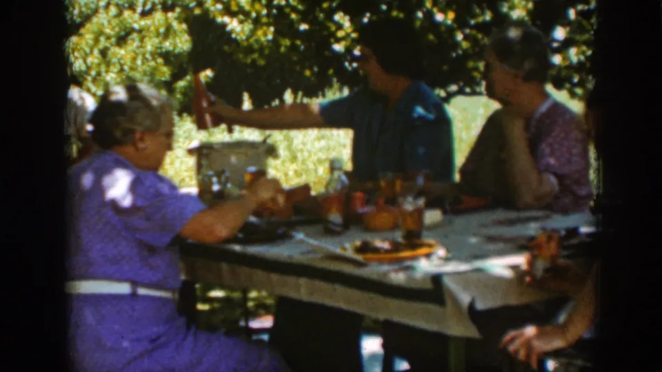 In the 1939 Auburn outdoor meal scene, adults sit beneath trees while a person in a blue shirt raises an item near the table.