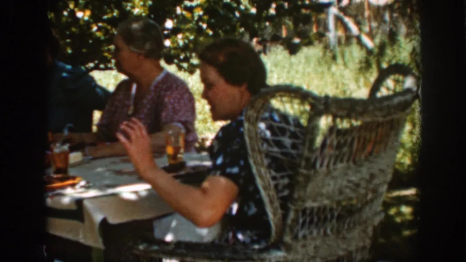 A 1939 Auburn outdoor family meal frame with adults seated near wicker chairs and amber glasses.