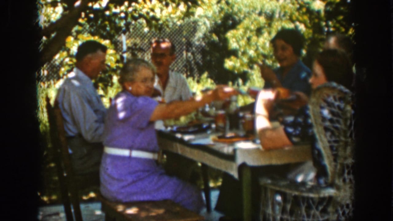 Auburn, California 1939 home-movie frame showing adults gathered at a shaded picnic table while an older woman in a purple dress serves food.