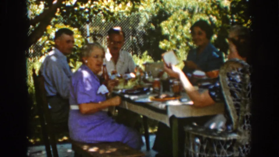 Auburn, California 1939 frame of a family picnic lunch with several adults seated at a table under trees, with dishes and drinks visible.