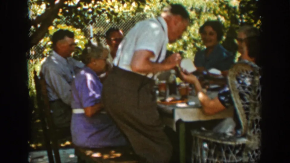 Auburn, California 1939 home-movie frame with a man in suspenders leaning into a family picnic table as seated adults watch or continue eating.