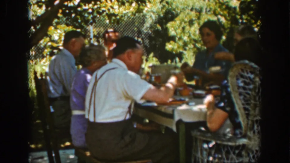 A man wearing suspenders sits in the foreground as adults share an outdoor meal in a leafy Auburn garden setting.