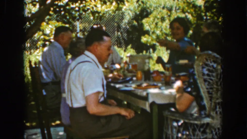 At the end of the clip, adults remain around the Auburn garden lunch table while a woman appears to gesture during conversation.