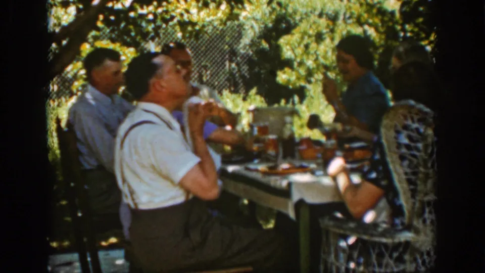 Auburn, California 1939 frame of adults dining outdoors as a man in suspenders lifts his hand near his face at the table.
