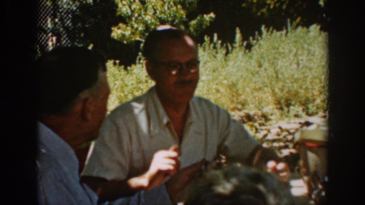 Two older men sit outdoors, one facing camera in glasses and a light shirt.