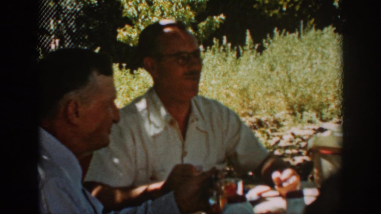 A man in glasses talks at an outdoor table while another man is seen in profile.