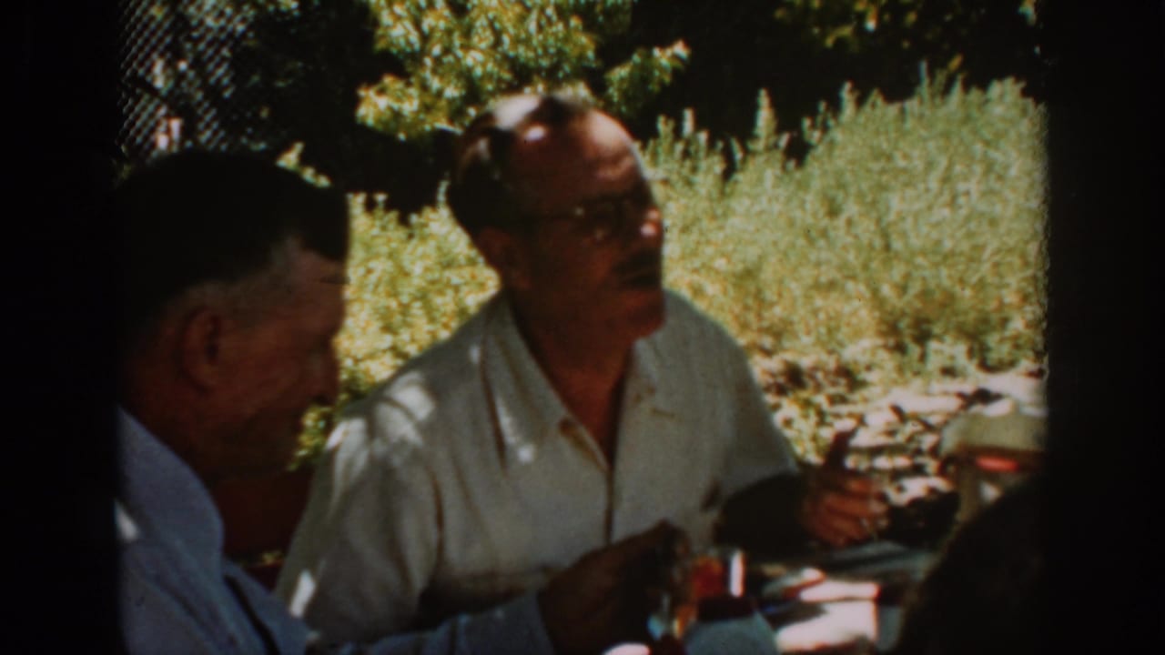 Two adult men seated outdoors at a shaded meal table.