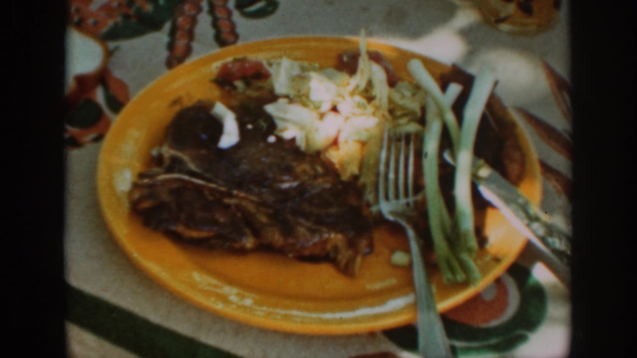 Yellow plate with grilled steak, coleslaw, green onions, and utensils on a patterned tablecloth.