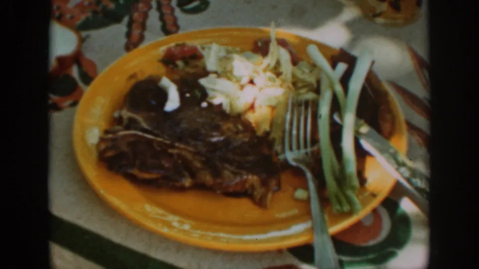 Auburn, California 1939 home-movie frame showing a steak meal on a yellow plate with side dishes and utensils outdoors.