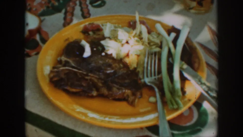 1939 Auburn home-movie close-up of a steak plate with coleslaw, green onions, fork, and knife in outdoor light.