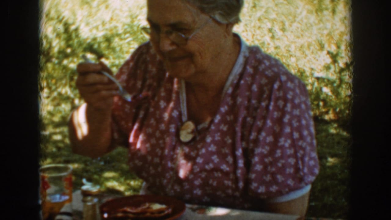 Older woman in glasses sits outdoors at a picnic table holding a spoon.