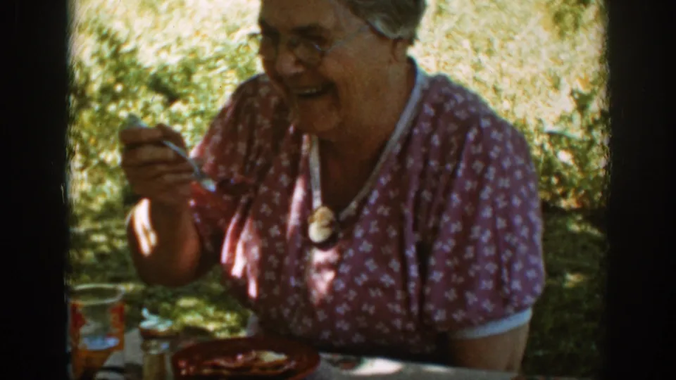 The older woman in a patterned dress smiles broadly beside picnic plates and glasses in the sun-dappled Auburn setting.