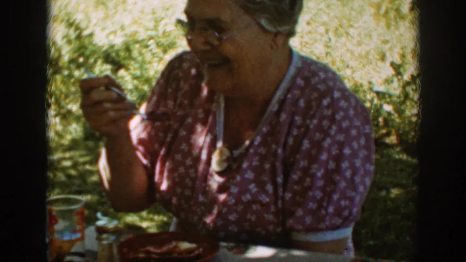 Smiling older woman in a floral dress holds a fork during an outdoor meal in Auburn, California, 1939.