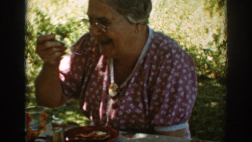 Older woman smiles as she lifts a fork above a plate at an outdoor lunch table in Auburn, California, 1939.