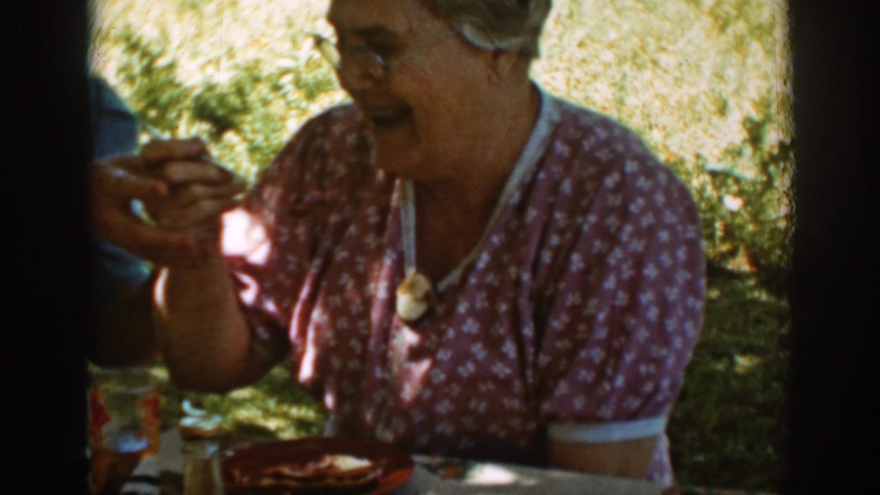 Older woman in a patterned blouse laughs while eating outdoors beside another adult.