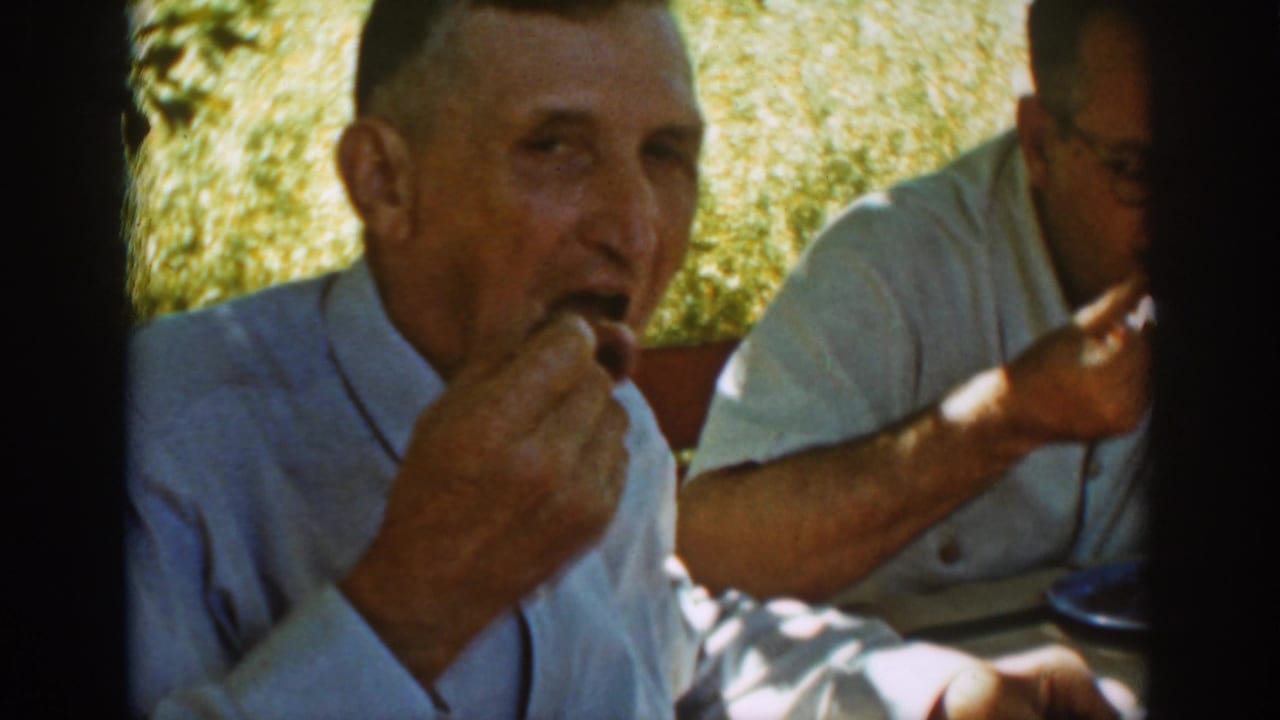 Man in a light shirt eats at an outdoor table with another adult partly visible nearby.