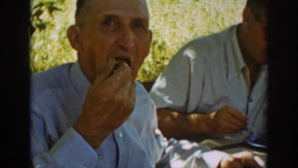 The man faces the camera while eating at a shaded outdoor table, with another seated adult partly visible beside him.