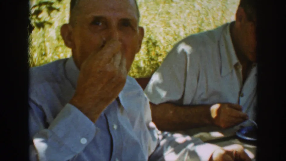 At the outdoor family picnic, the seated man raises his hand to his mouth while another adult sits beside him in partial view.
