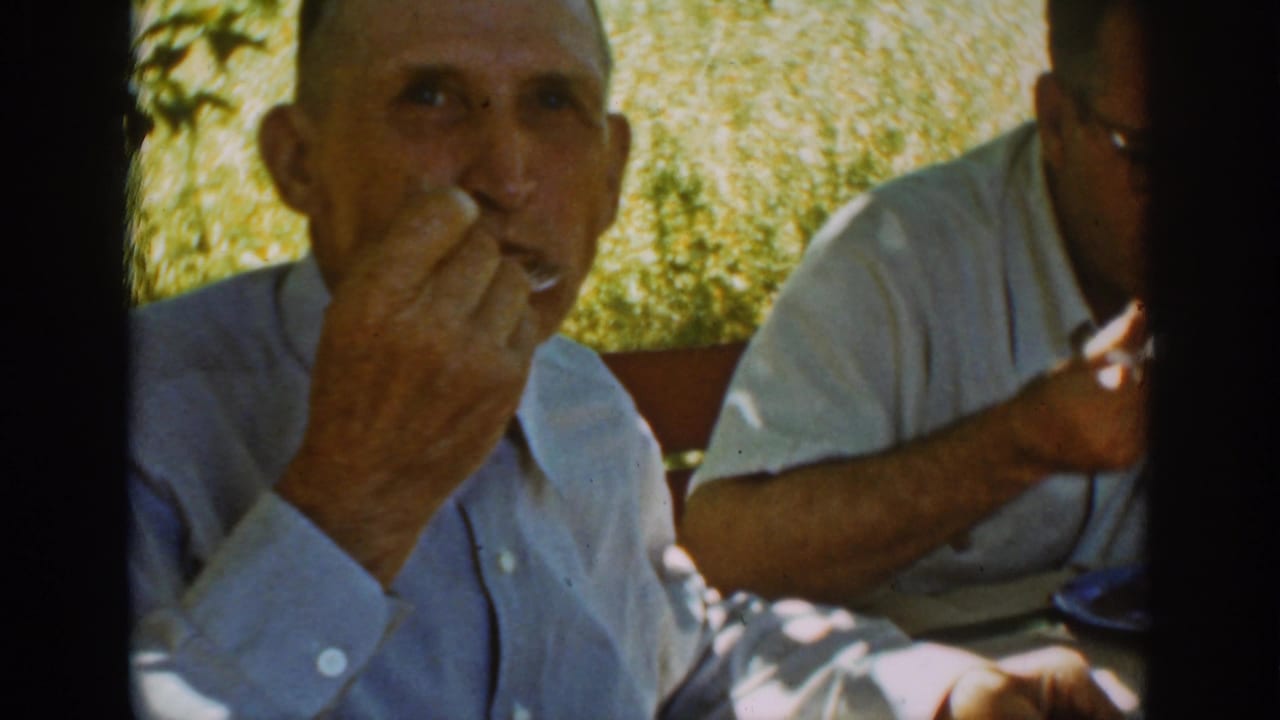 Two adult men sit outdoors at a meal, with the foreground man raising a fork to his mouth.