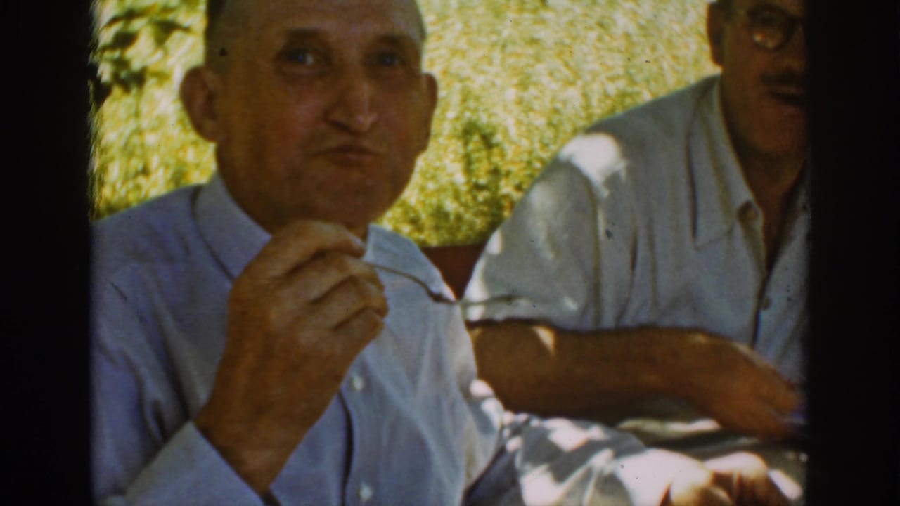Foreground man holds a fork while seated outdoors, with another man beside him at the table.
