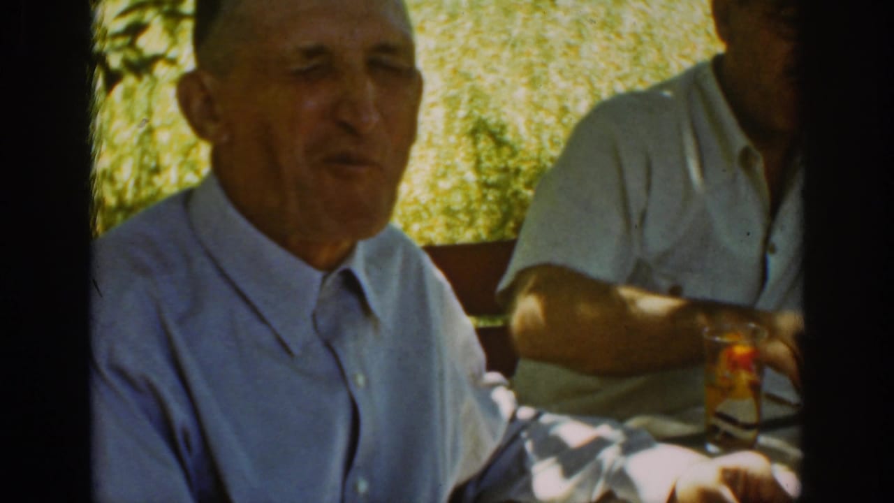 Two older men seated outdoors at a table in dappled sunlight.